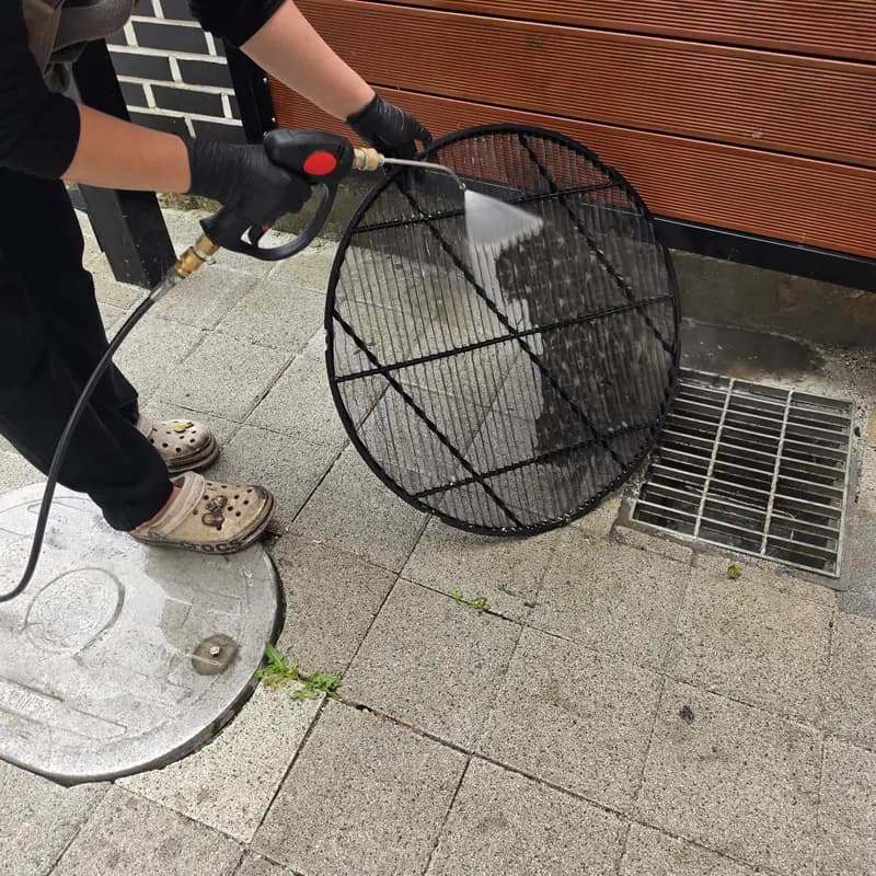 작업자가 실외 배수구 옆에서 분리한 원형 팬 보호망을 고압수로 헹구는 모습