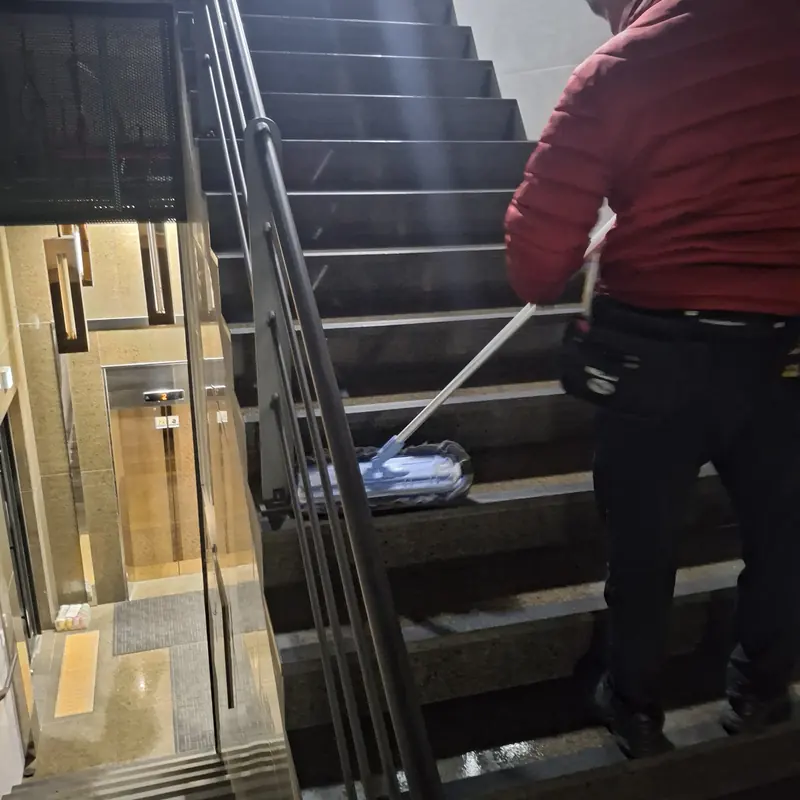 작업자가 건물 계단 디딤판을 밀대걸레로 닦고 난간 너머로 로비가 보이는 모습