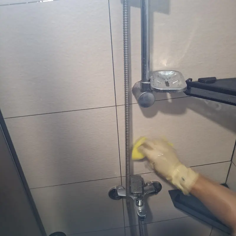 장갑을 낀 손이 노란 스펀지로 샤워부스 벽면 타일과 수전 주변을 닦는 모습