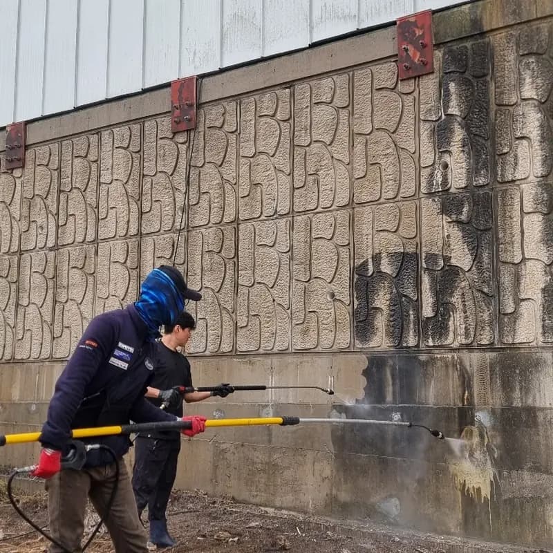 두 작업자가 연장 랜스가 달린 고압세척기로 외벽 하단 구간을 씻어내는 모습